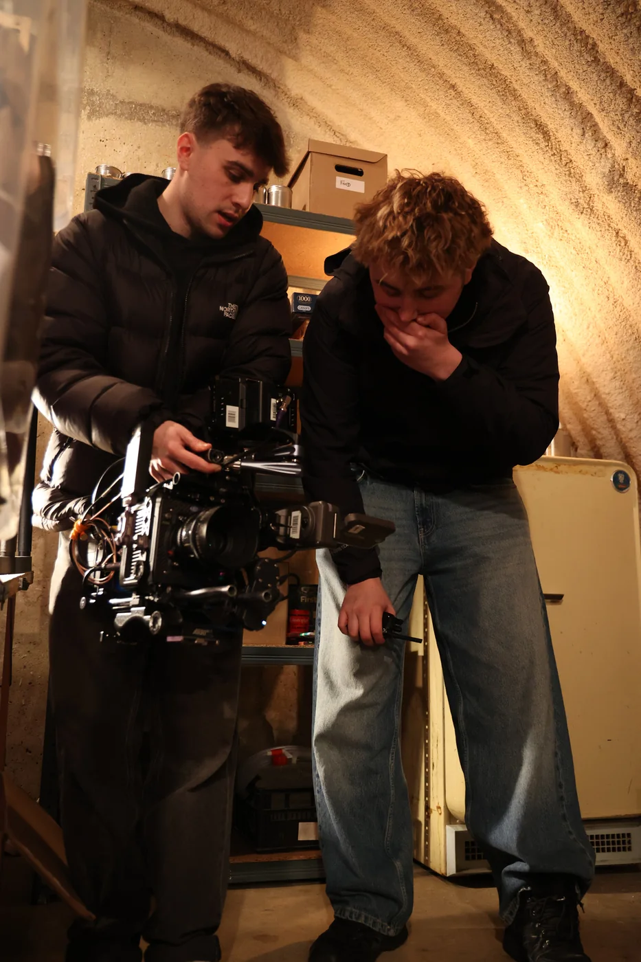 Two young crew members lean over a camera rig against the curved corrugated wall of a bunker